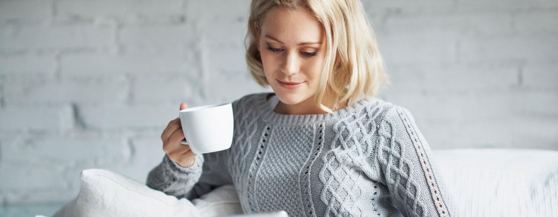 a woman sits on a couch with a cup of coffee and looks at her tablet